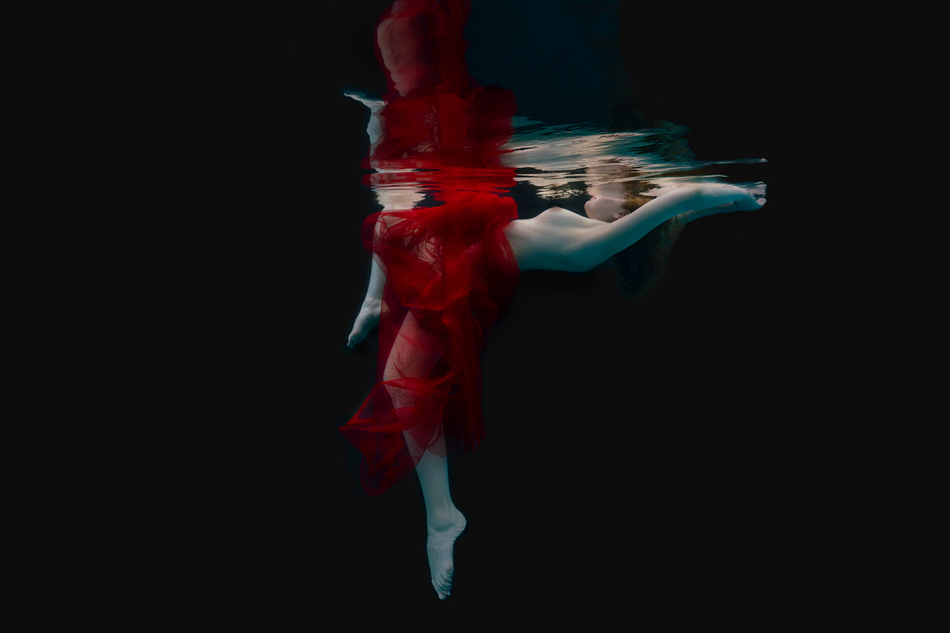 Within the depths of the sea, a stunning figure, wrapped in sheer red chiffon, floats gracefully amidst the aquatic realm. Her ethereal beauty and serene presence are accentuated by the play of light and shadow in the fine art photography. The mysterious allure of the underwater world surrounds her, evoking a sense of enchantment and tranquility