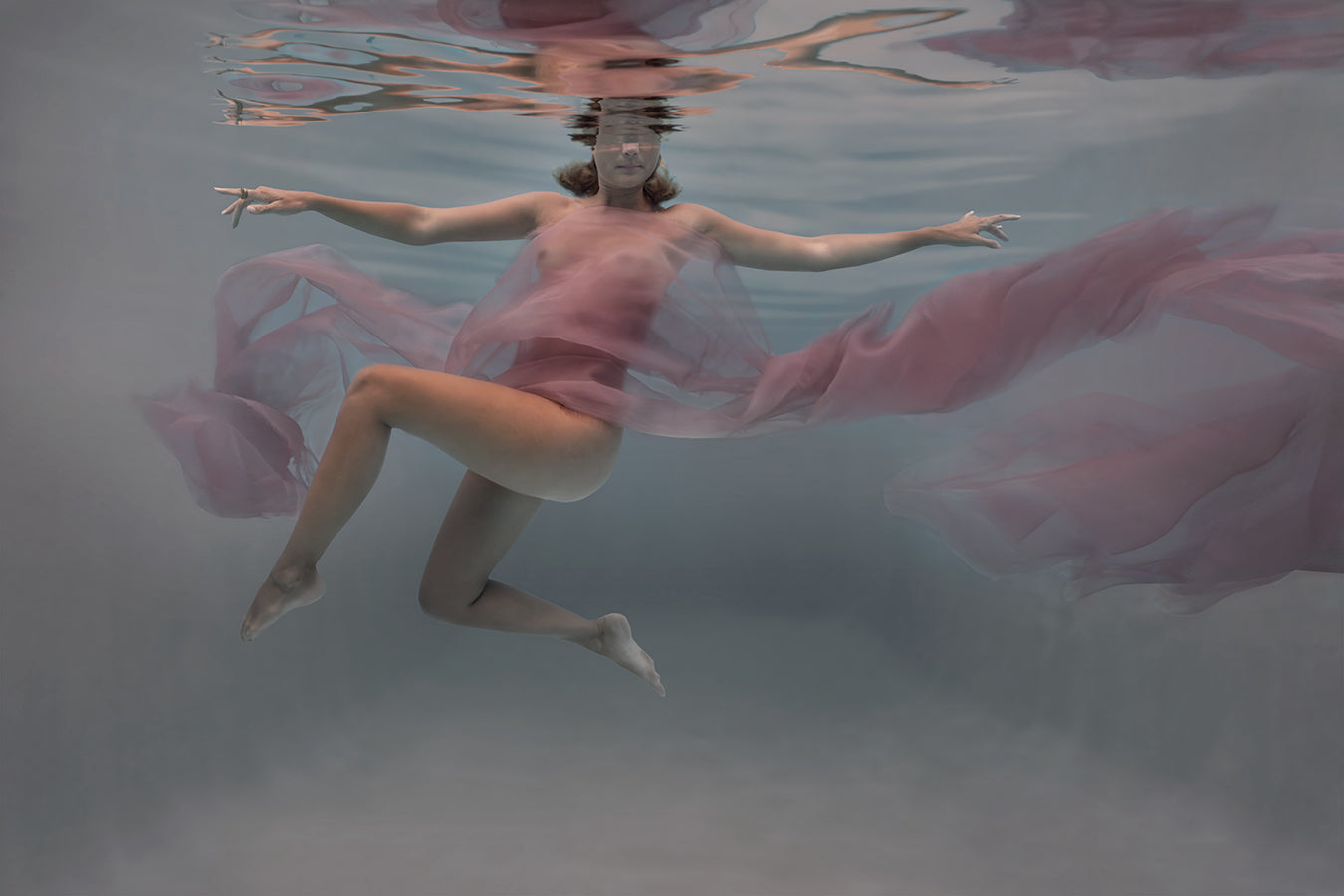 Within the depths of the sea, a stunning  figure, wrapped in sheer pink chifon, floats gracefully amidst the aquatic realm. Her ethereal beauty and serene presence are accentuated by the play of light and shadow in the fine art photography. The mysterious allure of the underwater world surrounds her, evoking a sense of enchantment and tranquility."